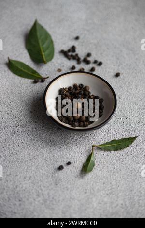 Eine Schüssel mit aromatischen schwarzen Pfefferkörnern, begleitet von frischen Lorbeerblättern auf einem strukturierten grauen Hintergrund. Stockfoto