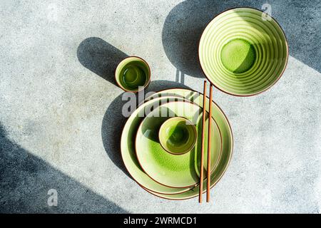 Blick von oben auf einen stilvollen Tisch mit grünem Keramikgeschirr, Schüsseln und einer Tasse mit braunen Essstäbchen, die alle in natürliches Sonnenlicht und Schatten getaucht sind Stockfoto