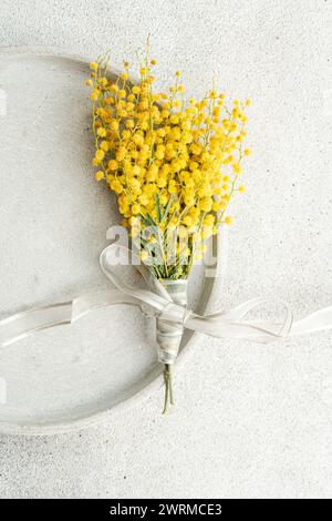 Blick von oben auf das zarte Arrangement von leuchtenden Mimosa-Blüten, die mit einem silbernen Band gebunden sind, präsentiert auf einer eleganten, strukturierten Tischoberfläche für einen eleganten Look Stockfoto