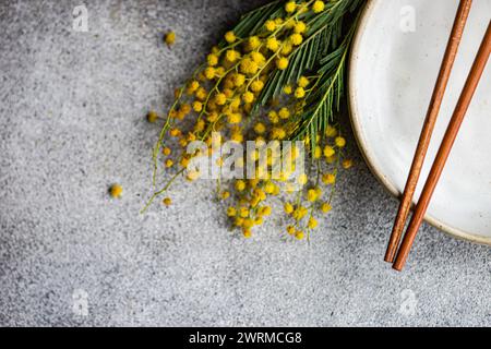 Eine detaillierte Ansicht der zarten Mimosa-Blüten neben Keramikgeschirr mit eleganten, kupfernen Essstäbchen auf strukturierter grauer Oberfläche Stockfoto