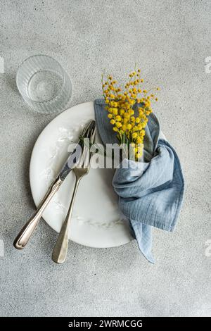 Elegantes Ambiente mit einem weißen, verzierten Teller, Vintage-Besteck, einer blauen Leinenserviette und einem leuchtend gelben Mimosa-Bouquet auf einem strukturierten Backdro Stockfoto