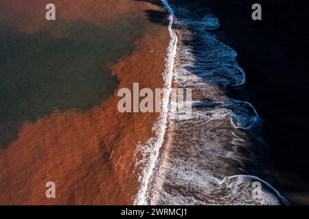 Luftaufnahme des starken Kontrasts zwischen dem rötlichen, mit Eisenoxid befleckten Wasser und dem unberührten weißen Schaum der Wellen am Strand Llumeres in Asturien, an Stockfoto
