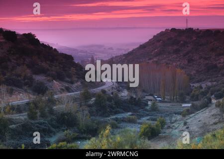 Ruhige Landschaft bei Sonnenaufgang mit einer gewundenen Straße, die durch Hügel und nebelige Täler unter einem lila Himmel führt Stockfoto