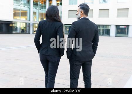 Rückansicht eines Mannes und einer Frau in professioneller Kleidung, die zusammen mit einem modernen Bürogebäude im Hintergrund laufen, was auf einen geschäftlichen Kontext hindeutet. Stockfoto