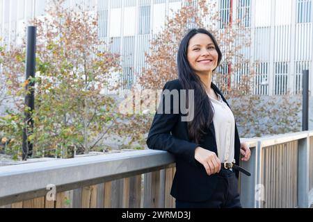 Lächelnde professionelle Frau in Geschäftskleidung, die vor einem modernen Bürogebäude steht. Stockfoto