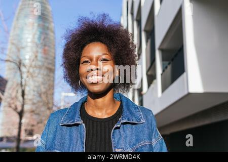 Eine strahlende afroamerikanische Frau mit einem selbstbewussten Lächeln steht in einer urbanen Landschaft, deren Architektur ihre natürliche Schönheit umrahmt Stockfoto