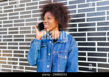 Eine afroamerikanische Frau genießt eine lebhafte Unterhaltung auf ihrem Telefon, ihr Lachen trägt zur urbanen Atmosphäre der Straßenlandschaft bei Stockfoto