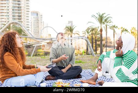 Eine vielfältige Gruppe von Freunden, darunter eine Afroamerikanerin im Hijab, genießt ein Picknick in einem Park mit Stadtgebäuden im Hintergrund. Stockfoto