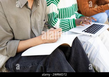Eine Gruppe von Schülern, die nicht erkennbar sind, die im Freien studiert werden, wobei einer im Notebook und der andere im Laptop schreibt. Stockfoto