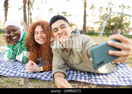 Multiethnische Gruppe von Studenten mit afroamerikanischer Frau, hispanischer Mann und kaukasischer Frau, die während eines Picknicks ein Selfie machen. Stockfoto