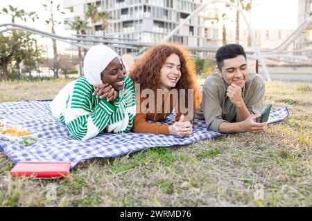 Drei lächelnde Studenten, die verschiedene Ethnien repräsentieren, darunter eine Frau in einem Hijab, die auf einer Picknickdecke liegt, wobei einer ein Smartphone hält Stockfoto
