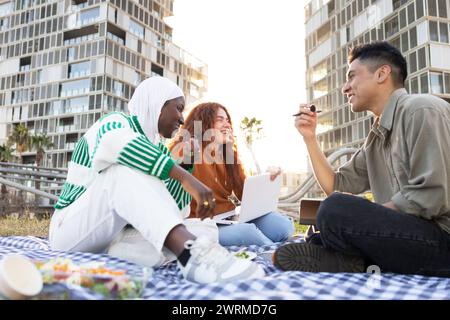 Verschiedene Gruppen von Studenten, darunter eine Afroamerikanerin und ein Mann, genießen ein Picknick und lernen mit einem Laptop an einem sonnigen Tag vor einem Stadtteil Stockfoto