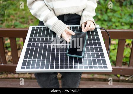 „Person im Freien, die ein Solarmodul zum Laden eines Smartphones nutzt, um einen nachhaltigen Energieverbrauch zu demonstrieren.“ Stockfoto