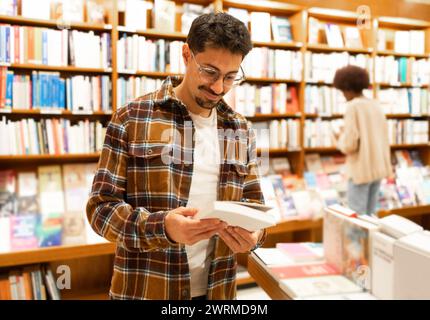 Ein junger Erwachsener in einem karierten Hemd wählt sorgfältig ein Buch aus einem Regal in einem gut beleuchteten Buchladen aus, umgeben von einer umfangreichen Sammlung. Stockfoto