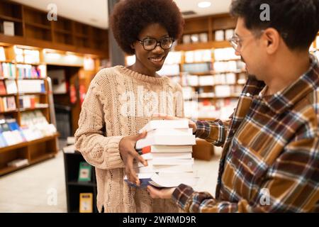 Zwei Freunde teilen einen fröhlichen Moment, während sie in einem gemütlichen Buchladen einen Stapel Bücher austauschen Stockfoto