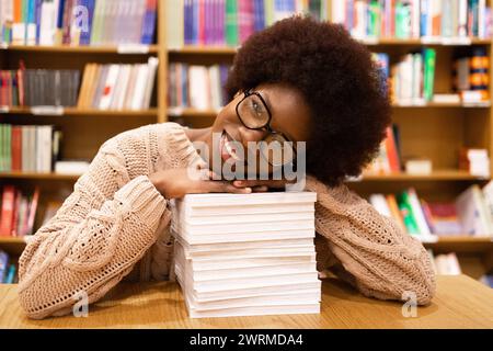 Eine fröhliche Afroamerikanerin legt ihren Kopf auf einen Stapel Bücher in einer Bibliothek, umgeben von Regalen mit farbenfroher Literatur Stockfoto
