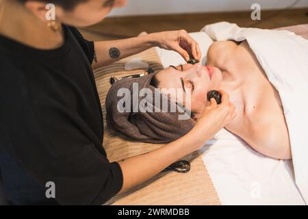 Eine ruhige Frau, die in einer ruhigen Spa-Umgebung eine Hot-Stone-Massage mit Fokus auf ihre Gesichtsmuskeln von einem professionellen Therapeuten genießt Stockfoto
