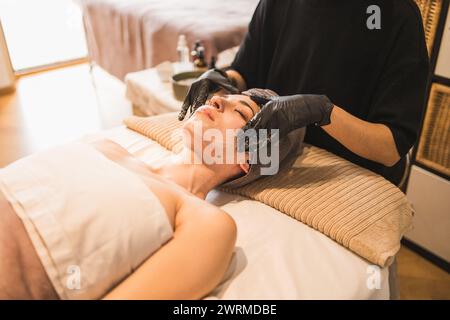 Eine ruhige Frau erhält eine pflegende Gesichtsmaske, die von einer Kosmetikerin in einer ruhigen Spa-Umgebung aufgetragen wird, um ihre Hautpflegeroutine zu verbessern Stockfoto