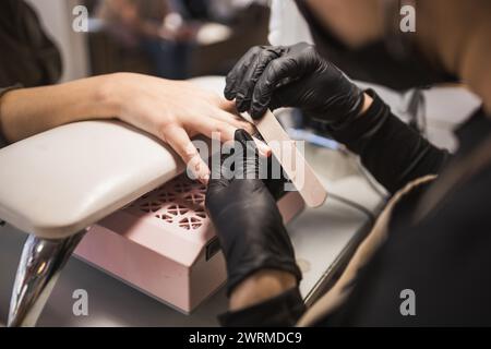 Ein erfahrener Manikürer in schwarzen Handschuhen feilt und formt die Nägel eines Kunden und konzentriert sich dabei auf Präzision und Pflege während einer Maniküre Stockfoto