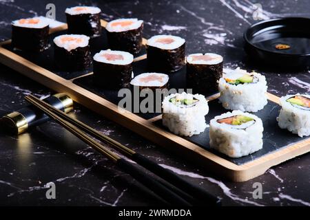 Eine elegante Auswahl an Sushi-Brötchen, die elegant auf einem Bambusteller präsentiert werden, ergänzt durch Essstäbchen und Sojasauce auf dunklem Marmorhintergrund. Stockfoto