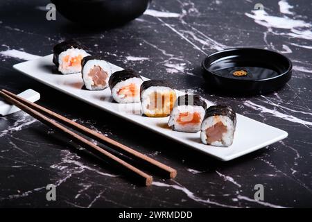 Verschiedene Sushi-Brötchen serviert auf einem weißen Teller mit Sojasauce und Essstäbchen auf dunklem Marmorhintergrund. Stockfoto