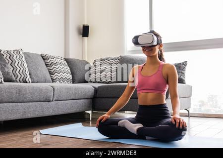Eine junge Erwachsene Frau asiatischer Herkunft lächelt, während sie in Sukhasana (lockere Pose) sitzt und ein Virtual-Reality-Headset in einem gut beleuchteten Wohnzimmer trägt Stockfoto