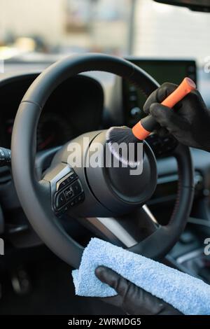 Nahaufnahme der Hände mit einer Bürste und einem Mikrofasertuch, das das Lenkrad in einem Fahrzeuginnenraum reinigt, wobei der Schwerpunkt auf die Fahrzeugwartung gelegt wird Stockfoto