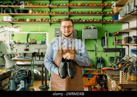 Ein fröhlicher Schuhmacher steht in einer gut ausgestatteten Werkstatt in Österreich und präsentiert ein Paar handgefertigter Lederschuhe mit farbenfrohen Schuhleisten. Stockfoto