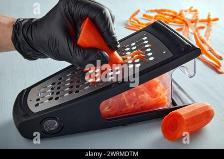 Kochen Sie in schwarzen Latexhandschuhen und zerkleinern Sie Karotten auf einer Metallreibe Stockfoto