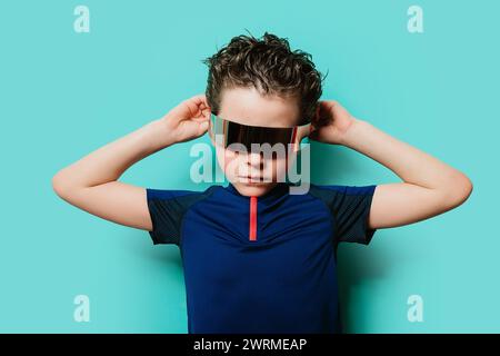 Ein trendiger kleiner Junge steht selbstbewusst mit Händen hinter dem Kopf und trägt ein futuristisches Visier vor türkisfarbenem Hintergrund Stockfoto