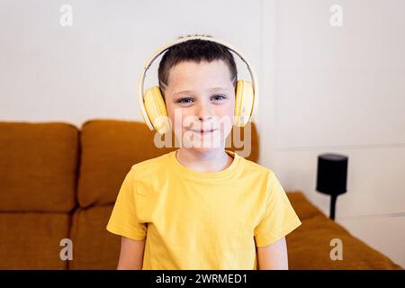 Ein kleiner Junge mit einem dezenten Lächeln, der ein hellgelbes T-Shirt und große gelbe Kopfhörer trägt, steht in einem Wohnzimmer Stockfoto