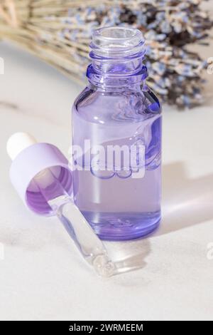 Eine elegante lavendelfarbene Glasflasche mit Pipettentropfen, im Hintergrund mit getrockneten Lavendelstielen, ideal für Aromatherapie oder kosmetische Anwendungen Stockfoto