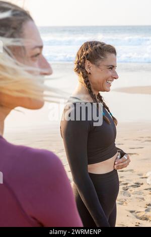 Zwei Frauen teilen einen freudigen Moment während eines friedlichen Yoga-Kurses an einem Sandstrand vor der Kulisse eines atemberaubenden Sonnenuntergangs. Stockfoto