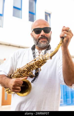 Ein bärtiger Mann in einer Sonnenbrille, der ein Saxophon hält, mit selbstbewusstem Ausdruck in die Kamera blickt, in lässiger Kleidung gekleidet, um Jazz Internati zu feiern Stockfoto