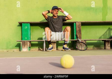 Man sieht einen Mann, der eine Pause von einem Tennisspiel macht, auf einer Bank mit den Händen auf dem Kopf ruht und eine Rückansicht zeigt. Stockfoto