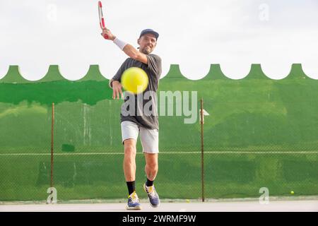 Ein Mann spielt aktiv Tennis auf einem Platz im Freien, während er einen Tennisball trifft, der von der Kamera weg zeigt. Stockfoto