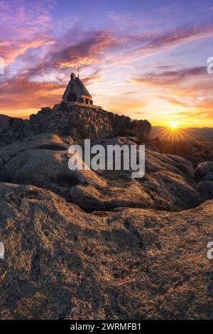 Ein ruhiger Sonnenuntergang wirft warmes Licht über eine einzigartige Kapelle, die auf zerklüfteten Bergfelsen vor einem dramatischen Himmel thront. Stockfoto