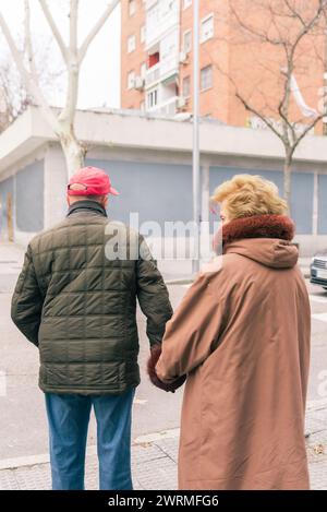 Ein älteres Ehepaar geht Hand in Hand, von hinten gesehen, auf einer Stadtstraße mit Gebäuden im Hintergrund, die die Gesellschaft zum Ausdruck bringen Stockfoto
