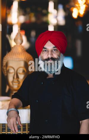 Der selbstbewusste Koch mit einem roten Turban steht in einem Restaurant mit einer Buddha-Statue im Hintergrund Stockfoto