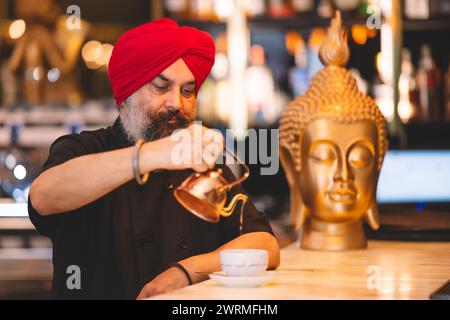 Ein Sikh-Mann mit rotem Turban und Bart gießt vorsichtig Kaffee in einem Café, mit einer Buddha-Statue im Hintergrund, was die kulturelle Vielfalt veranschaulicht Stockfoto