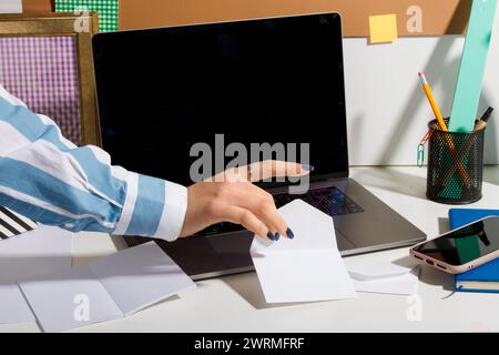 Abgeschnittene, nicht erkennbare weibliche Büroangestellte, die auf einem gut organisierten Schreibtisch mit verschiedenen Büromaterialien neben einem Laptop eine Notiz schnappt. Stockfoto