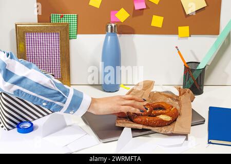 Eine zugeschnittene, nicht erkennbare Frau genießt eine Brezel während einer Mittagspause an einem überladenen Bürotisch mit einem Laptop, Haftnotizen und einer wiederverwendbaren Wasserflasche Stockfoto