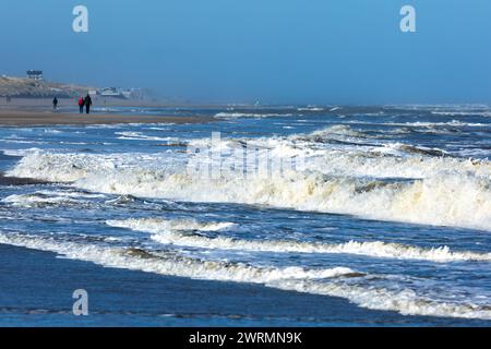 Egmond aan Zee, Niederlande - 17. März 2022: Wellen in der rauen Nordsee mit einigen Menschen, die in der Sonne am Ufer entlang laufen Stockfoto