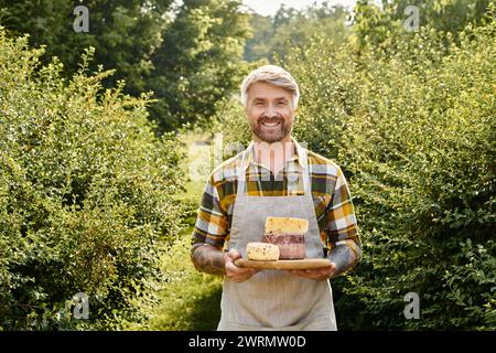 Fröhlicher gutaussehender Bauer mit Tattoos, der hausgemachten Käse in der Hand hält und in die Kamera lächelt Stockfoto