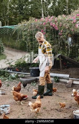 Gut aussehender, fröhlicher Mann mit Tattoos, der Hühner in ihrer Voliere füttert, während er auf seiner Farm im Dorf ist Stockfoto