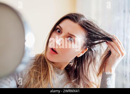 Die junge schockierte Frau ist überrascht zu sehen, wie schmutziges, fettiges Haar im Spiegel zu Hause aussieht. Gesundheitsbehandlung. Haarpflegeprodukte. Selbstpflege Stockfoto