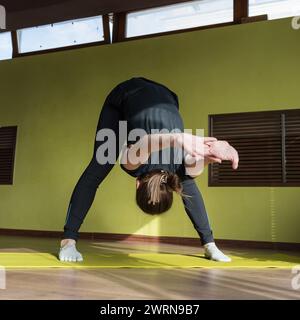 Athletic Young Woman führt Dandayamana Mudrasana-Trainingsvarianten durch, neigt sich nach vorne mit zurückgewendeten Armen, trainiert während er auf der Matte steht Stockfoto