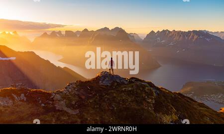 Männlicher Wanderer genießt das Labyrinth der Mitternachtssonne auf dem Gipfel des Berges in reine, Lofoten, Norwegen Stockfoto