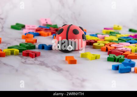 Schöner kleiner Marienkäfer aus hellem Modellierton, der auf weißem Marmortisch zwischen verschiedenen kleinen farbigen Holzbuchstaben steht. Stockfoto
