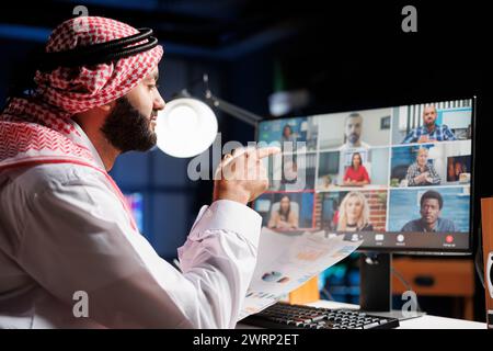 Seriöser arabischer Geschäftsmann, der Papiere hält und Ideen mit seinem Team über ein Videogespräch auf dem Desktop-pc diskutiert. Muslimischer Kerl, der Informationen mit Kollegen in einem Online-Meeting vergleicht. Stockfoto